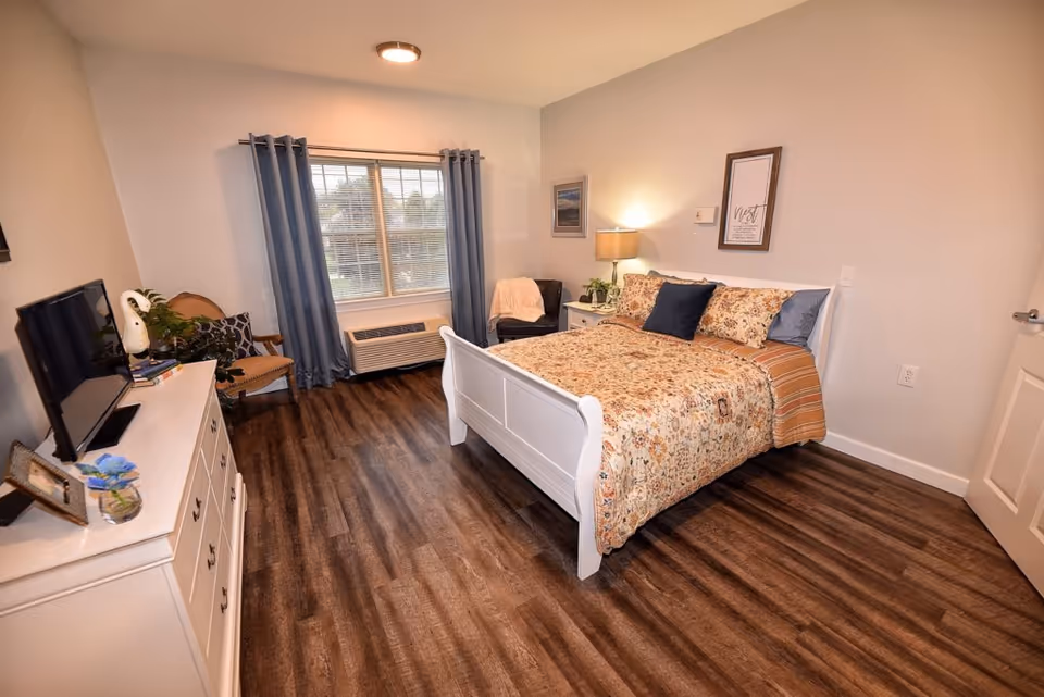 Furnished bedroom with a white bed, dresser and TV, and a chair by a window with blue curtains.