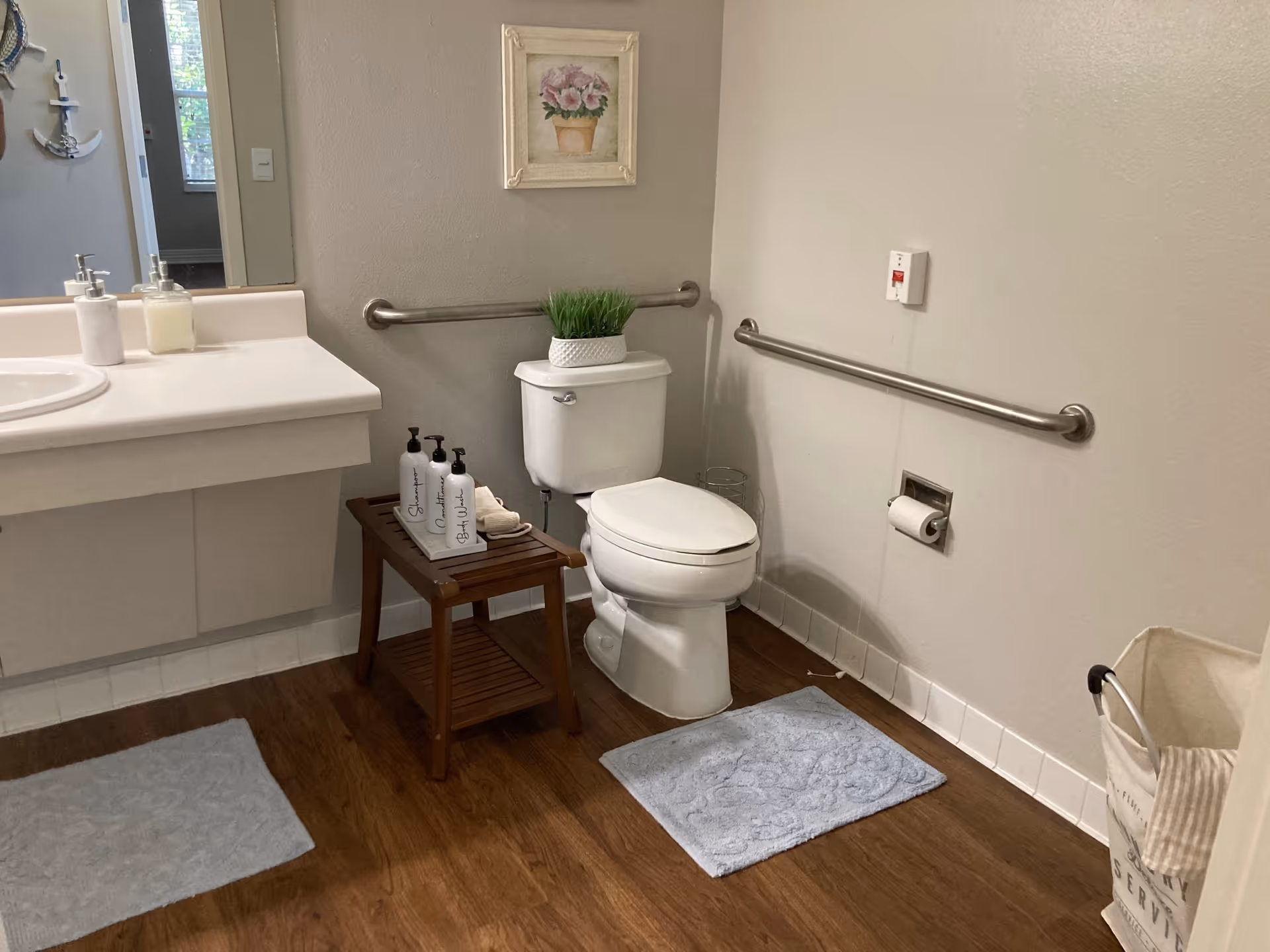 A clean and accessible bathroom with a white toilet, grab bars on the walls, a wooden stool holding three bottles and a towel, a white sink countertop with soap dispensers, two light blue bath mats on a wooden floor, a framed floral picture on the wall, and a laundry basket in the corner.