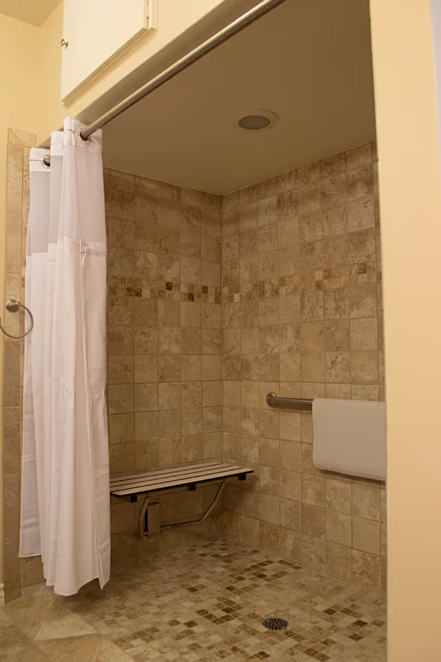 A tiled walk-in shower with beige and brown square tiles on the walls and floor. The shower has a white curtain on a metal rod, a fold-down bench attached to the wall, and a metal grab bar with a white towel hanging on it.