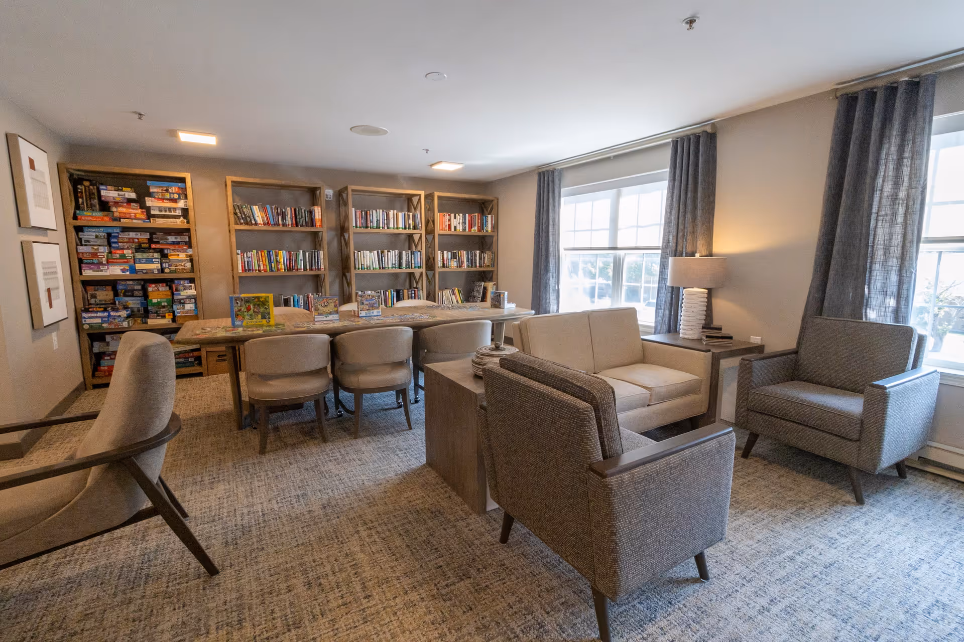 A cozy reading and game room with bookshelves filled with books and board games, a long table with chairs, and a seating area with a sofa and armchairs near large windows with gray curtains.