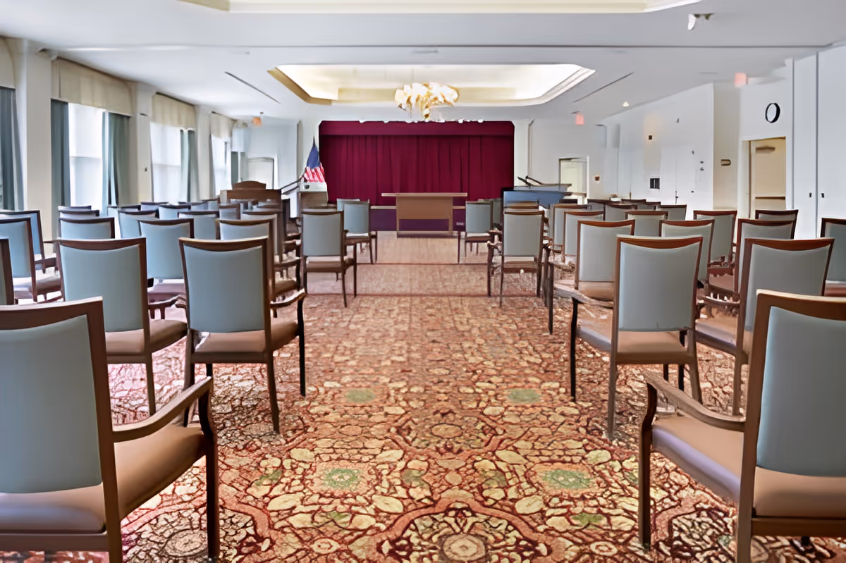 A large room with rows of chairs facing a stage with a podium and an American flag. The room has a patterned carpet, white walls, large windows with curtains on the left side, and a chandelier hanging from the ceiling.