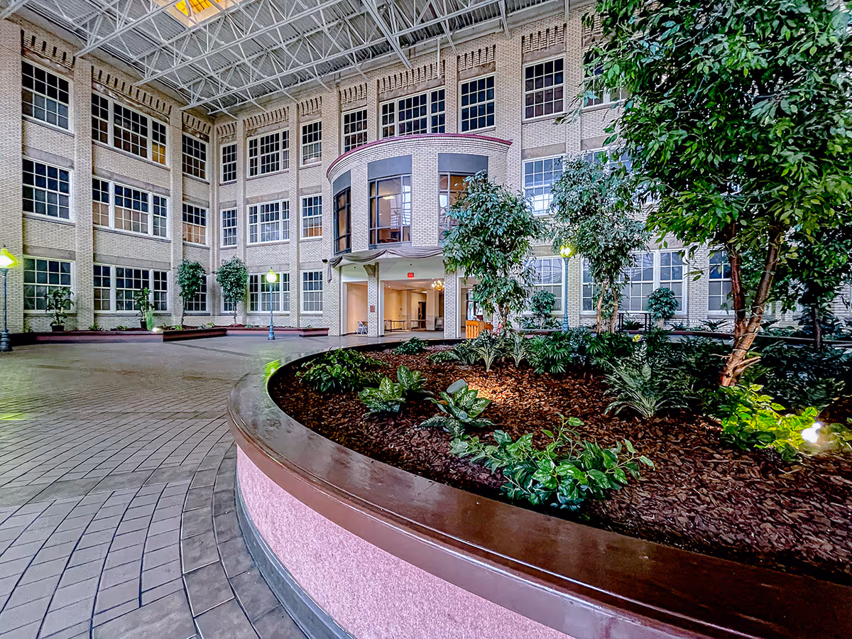 Indoor courtyard area of a senior living facility with a large planter filled with green plants and trees, surrounded by a tiled floor. The courtyard is enclosed by a multi-story brick building with many windows and a glass ceiling allowing natural light to enter. There are lamppost-style lights and an entrance to the building visible in the background.