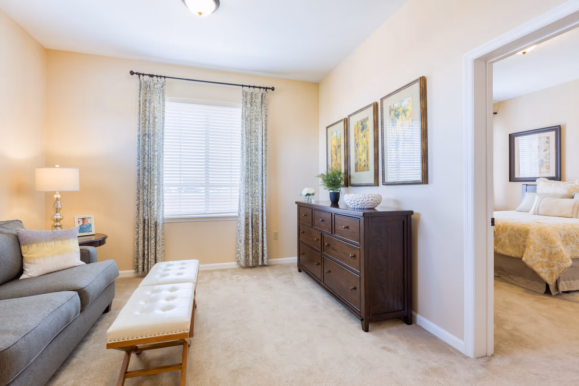 A cozy living room area with a gray sofa, a white cushioned bench, a side table with a lamp and picture frame, and a window with patterned curtains. On the right side, there is a dark wooden dresser with decorative items and three framed pictures above it. An open doorway leads to a bedroom with a bed covered in light-colored bedding and a framed picture on the wall.