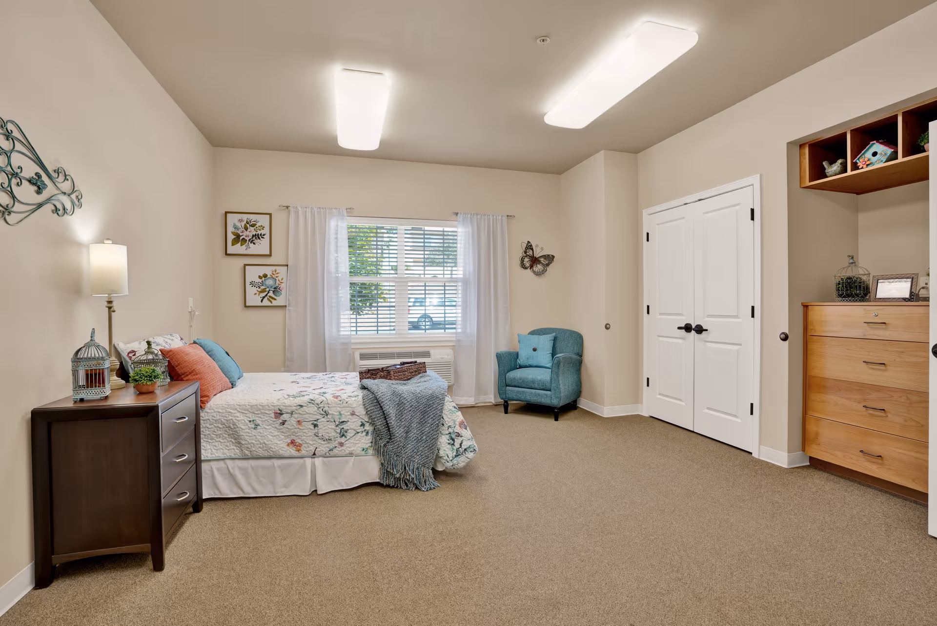 A cozy bedroom with a bed covered in floral bedding and colorful pillows, a dark wooden nightstand with a lamp and decorative items, a teal armchair near a window with white curtains, a double-door closet, and a wooden dresser with shelves above it. The room has beige walls and carpeted floor.