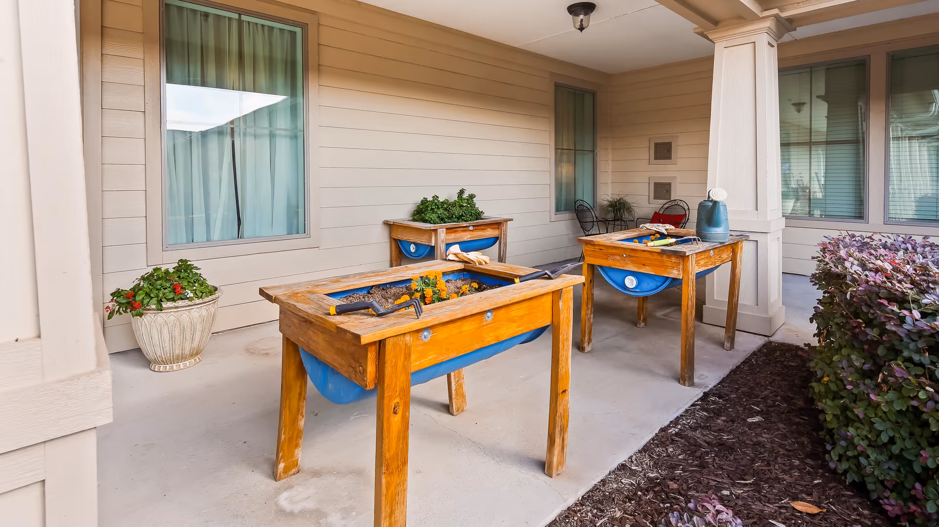 Covered outdoor patio area with three wooden raised garden beds containing soil and some plants. Gardening tools and a watering can are placed on the garden beds. There are windows and beige siding on the building wall, a potted plant on the left, and some shrubs on the right side.