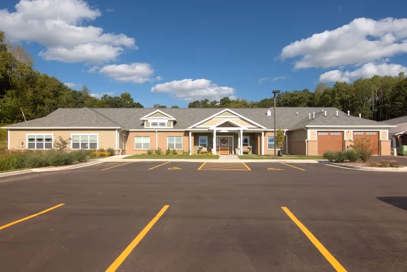 Front exterior view of a single-story senior living facility building with a covered entrance, surrounded by greenery and a parking lot with marked spaces in front under a partly cloudy sky.