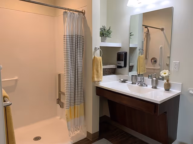 Accessible bathroom with a walk-in shower with grab bars and striped curtain, a vanity with sink and mirror, soap dispenser, and yellow towels.