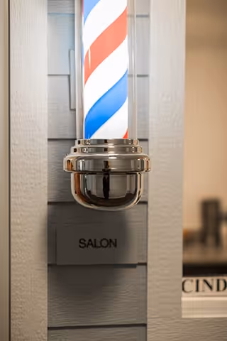 Close-up of a traditional barber pole with red, white, and blue stripes mounted on a gray wall next to a sign that reads 'SALON'.