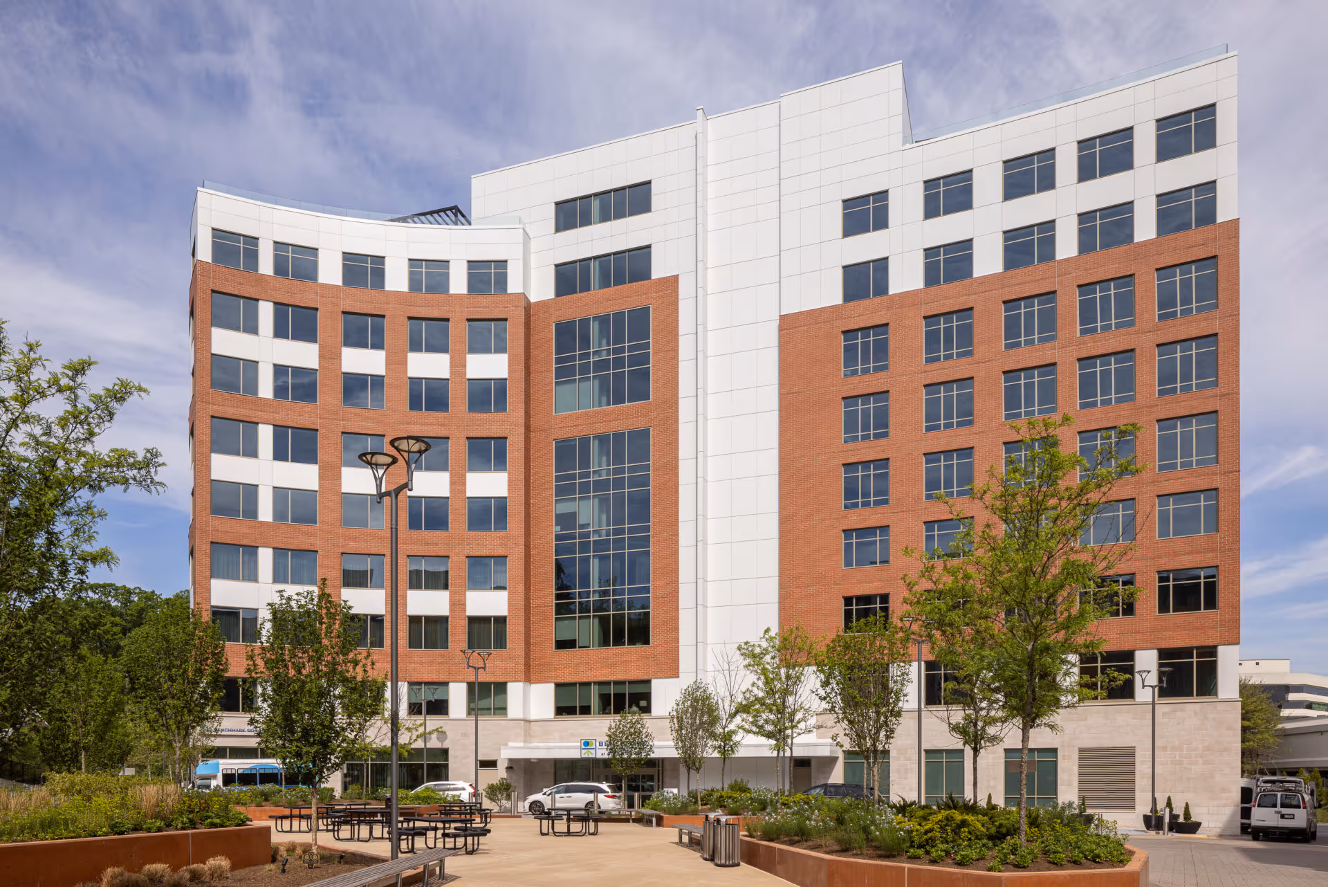 Exterior view of a modern multi-story building with a combination of red brick and white paneling. The building has many windows and is surrounded by landscaped greenery, trees, and outdoor seating areas with tables and benches under a partly cloudy sky.