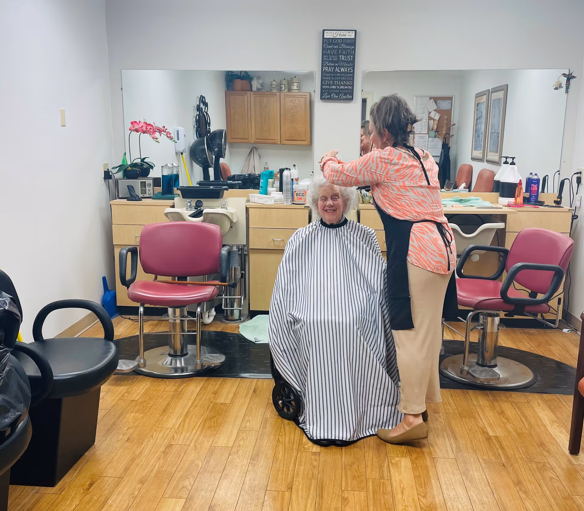An elderly woman with white hair is sitting in a wheelchair covered with a black and white striped cape, smiling while a hairstylist wearing a pink patterned top and beige pants is cutting her hair in a salon setting. The room has wooden flooring, salon chairs, a large mirror, and cabinets with various hair products.