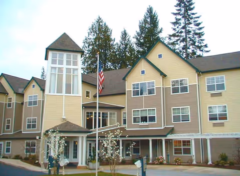 Exterior view of a multi-story senior living facility building with beige and brown siding, multiple windows, a prominent tower-like structure, an American flag on a flagpole in front, and some small trees and shrubs in the landscaped area.