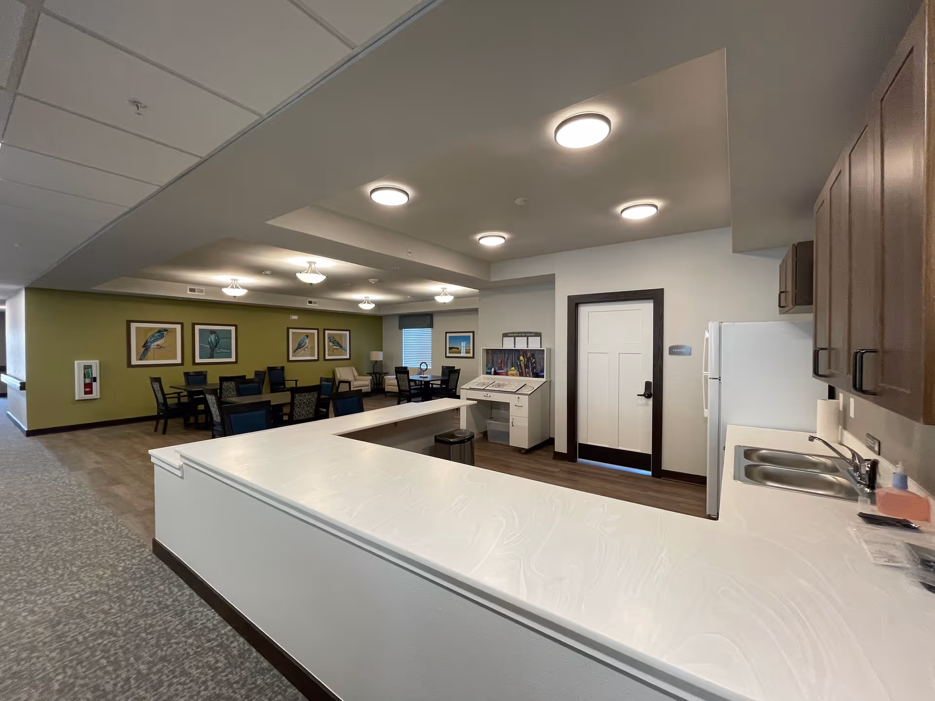 Interior view of a senior living facility common area with a kitchen counter in the foreground, a refrigerator, sink, and cabinets on the right, and a dining area with tables, chairs, and bird artwork on a green accent wall in the background.