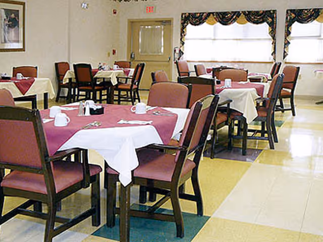 Dining room with multiple tables covered with white and maroon tablecloths, each set with cups, napkins, and utensils. The room has several chairs with maroon cushions, large windows with valances, and a door in the background.