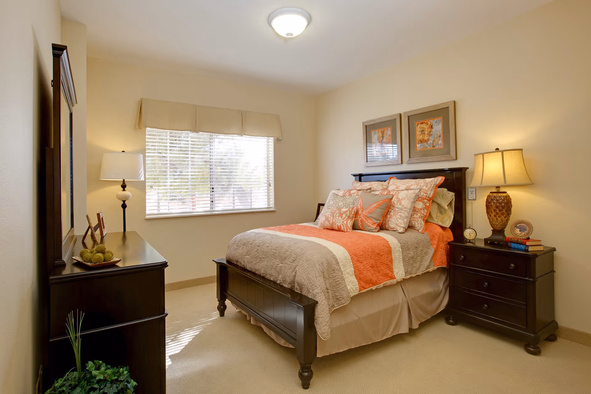 A cozy bedroom with a bed featuring a beige and orange quilt and multiple decorative pillows. There is a window with blinds and a beige valance letting in natural light. On one side of the bed is a dark wooden nightstand with a lamp, clock, books, and a framed photo. On the opposite side is a matching dark wooden dresser with a mirror, a lamp, and decorative items. The walls are painted light beige and the floor is carpeted.