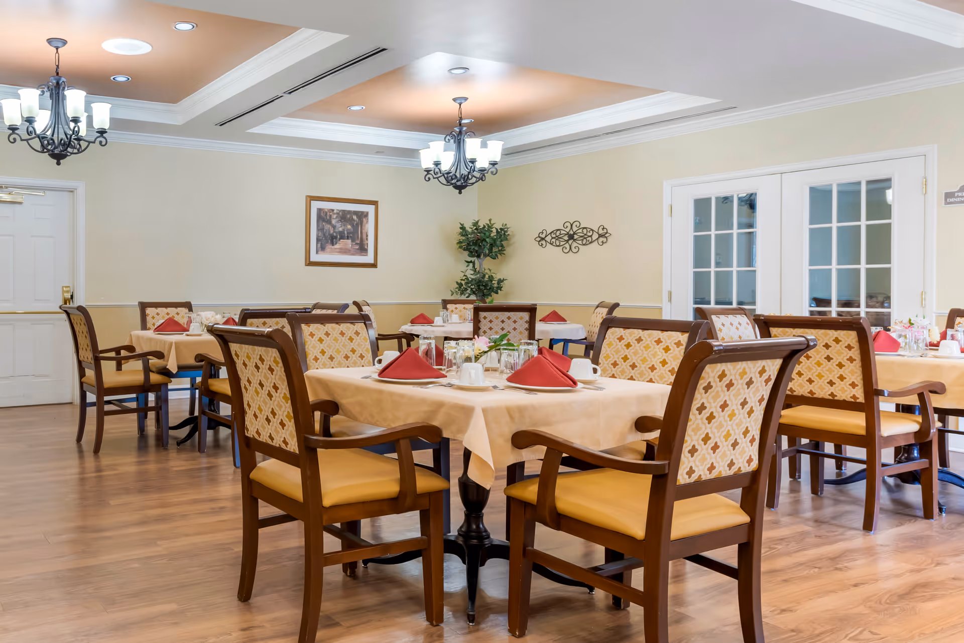 A dining room with several tables covered with beige tablecloths, each set with red folded napkins, white cups, glasses, and silverware. The chairs have wooden frames with patterned upholstery and yellow seat cushions. The room has wooden flooring, cream-colored walls, two chandeliers, a framed picture, a decorative wall piece, and a potted plant in the corner. There are double glass doors on one side of the room.
