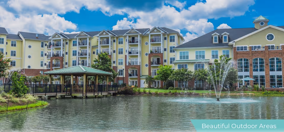 View of a large senior living facility building with multiple floors and balconies, situated behind a pond with a water fountain. There is a covered wooden gazebo on the edge of the pond, surrounded by landscaped greenery under a partly cloudy blue sky.