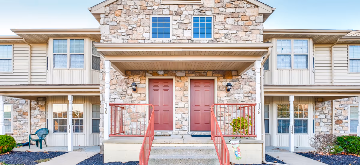 Front exterior view of a senior living facility building with stone and beige siding, featuring two red entrance doors with small porches and red railings, windows on both floors, and a clear blue sky.