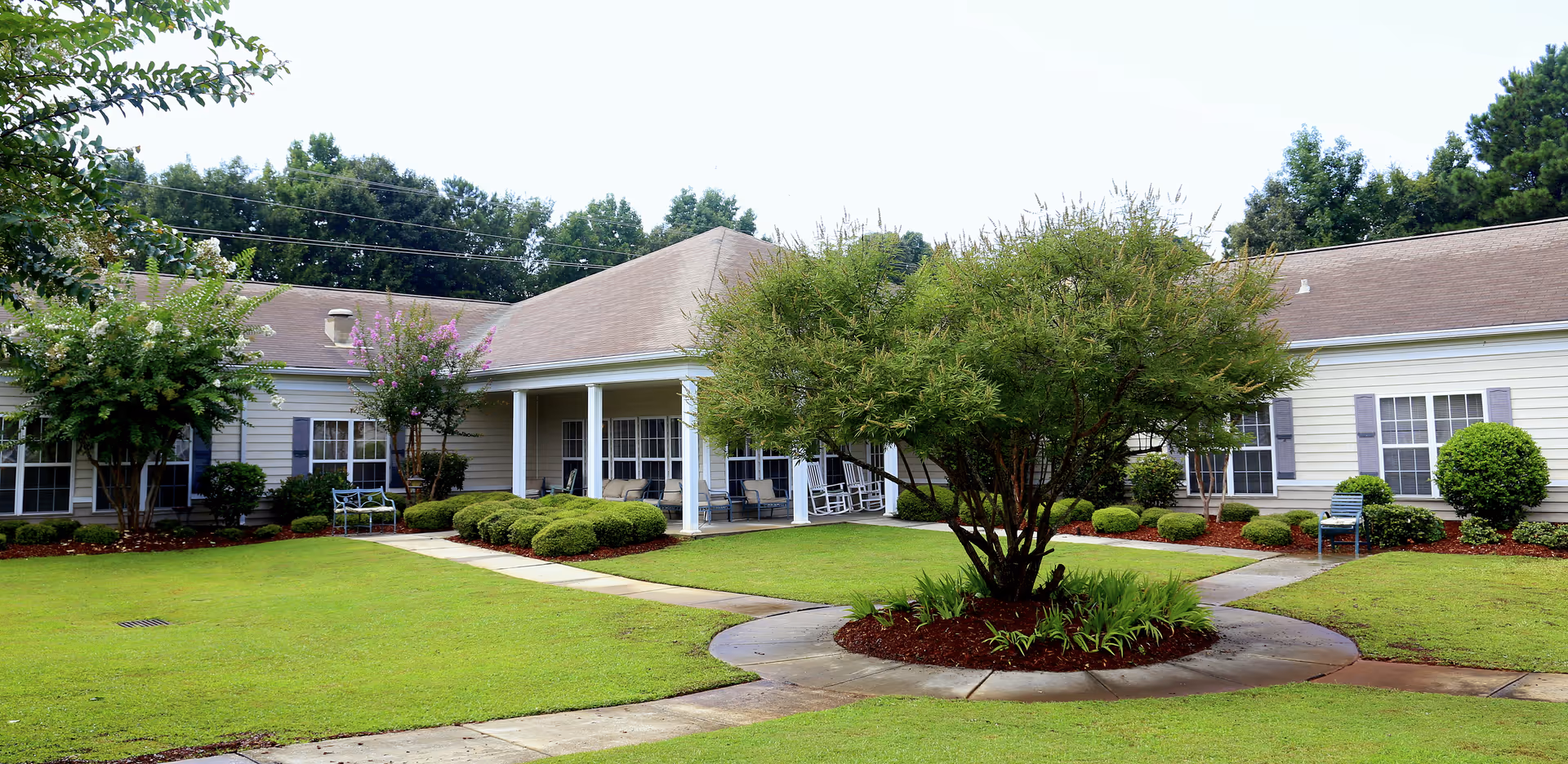 A single-story building with a beige exterior and a brown roof surrounding a green courtyard. The courtyard features a large tree in the center with a circular mulch bed, neatly trimmed bushes, and a well-maintained lawn. There are several windows with purple shutters and a covered porch area with chairs and rocking chairs. Trees and greenery are visible in the background under a clear sky.