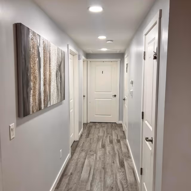 A clean, well-lit hallway with light gray walls and wood-patterned flooring. There are multiple white doors on both sides and one door at the end marked with the letter 'E'. A large abstract painting with neutral tones hangs on the left wall.