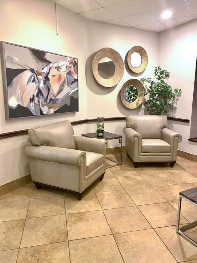 A cozy corner seating area with two beige leather armchairs separated by a small dark wooden side table. The walls are decorated with three round mirrors with textured frames and an abstract painting. A green leafy plant is placed in the corner, and the floor is tiled with large beige tiles.