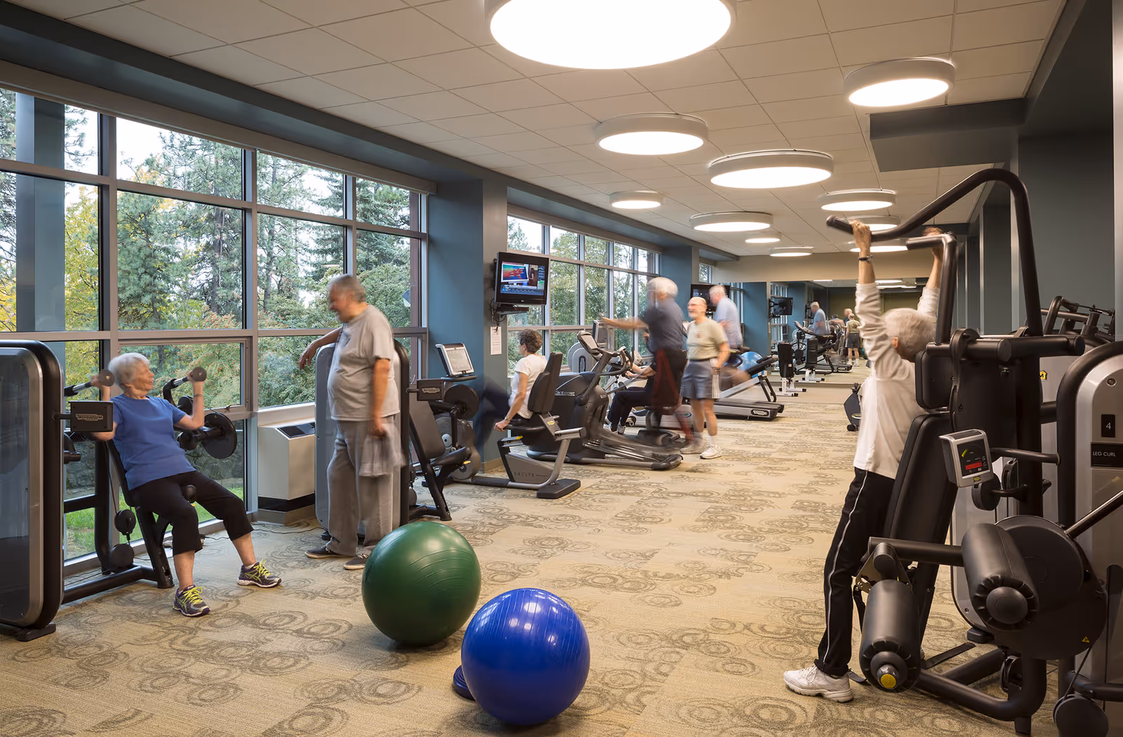 A fitness room with large windows showing trees outside. Several elderly people are using various exercise machines and equipment, including weight machines, stationary bikes, and exercise balls. The room has circular ceiling lights and carpeted flooring with a patterned design.