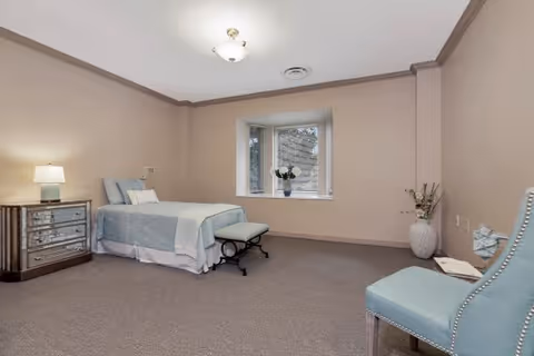 A simple, clean bedroom with a single bed covered in light blue bedding, a small bench at the foot of the bed, a mirrored nightstand with a lamp, a window with a potted plant on the sill, and a light blue upholstered chair with a book on it. The walls are beige and the carpet is gray.