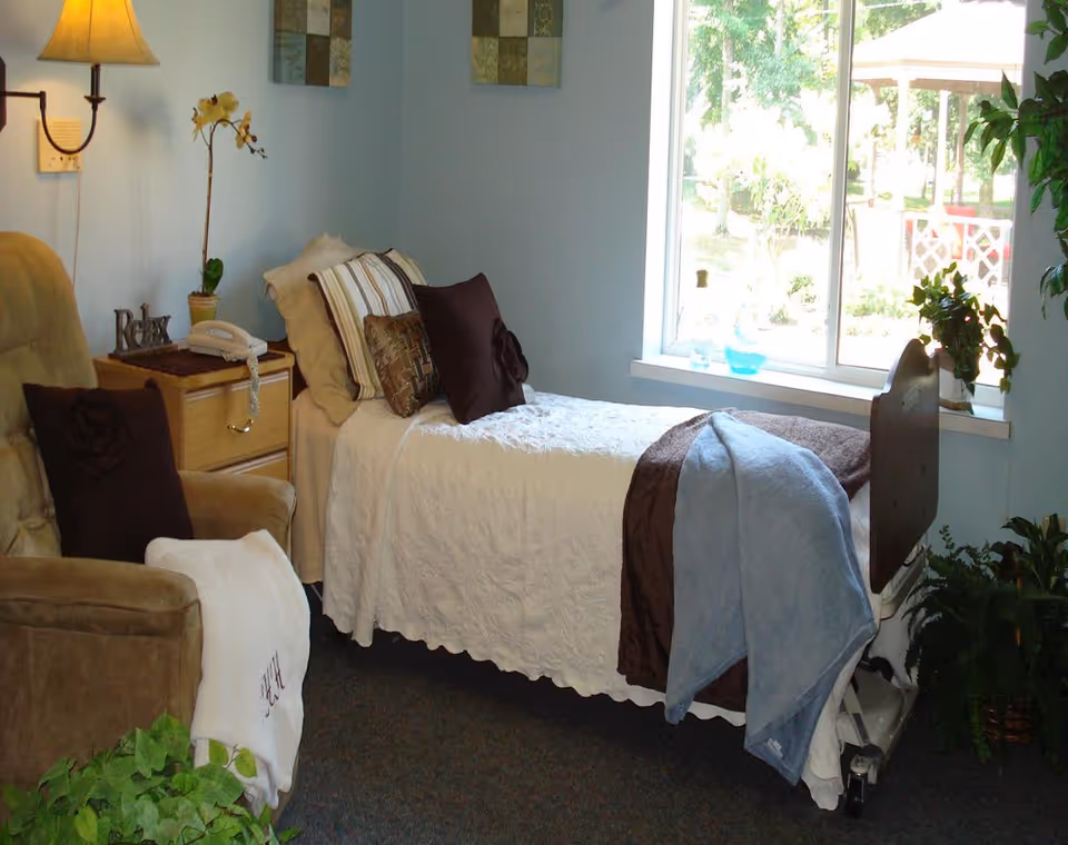 A cozy single bedroom with a bed, recliner, nightstand and a window overlooking a garden.
