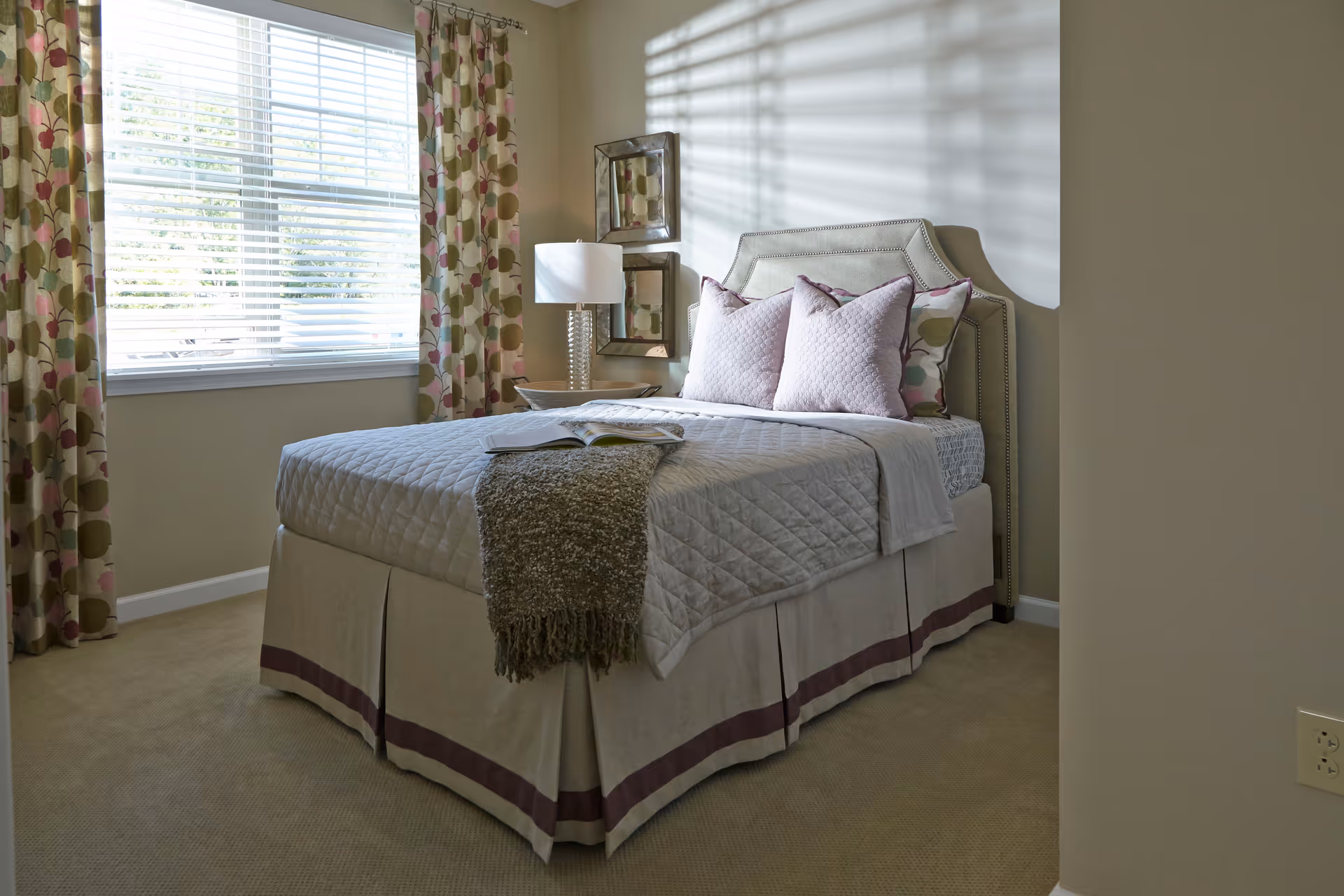 A neatly made bed with a quilted bedspread, three decorative pillows, and a throw blanket in a bedroom with beige walls and carpet. There is a window with floral curtains allowing natural light to enter the room. A bedside table with a lamp and two square mirrors on the wall are visible.