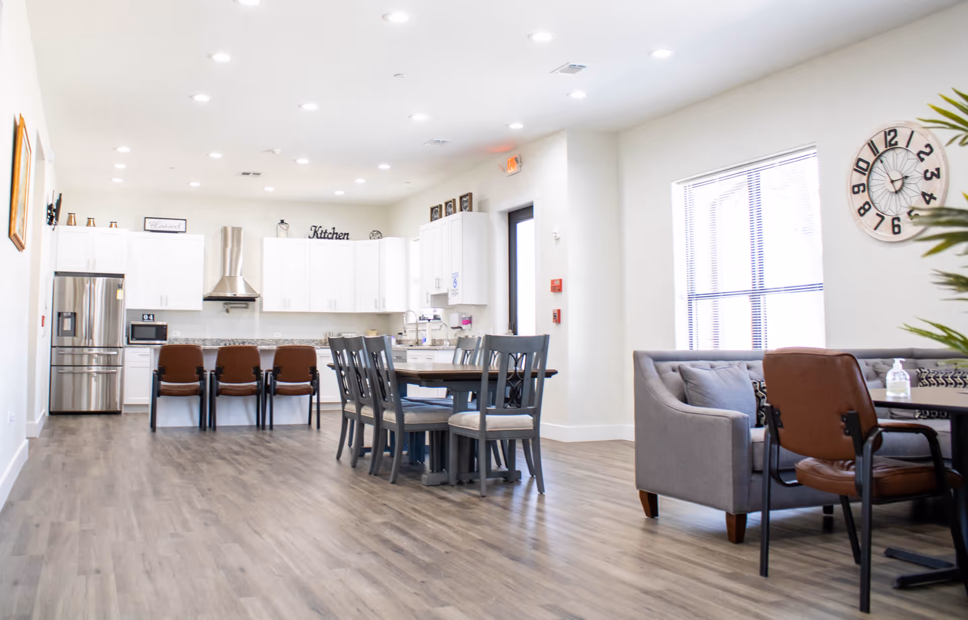 Bright and spacious assisted living facility kitchen and dining area with white cabinets, stainless steel refrigerator, a kitchen island with brown chairs, a dining table with gray chairs, and a seating area with a gray couch and brown chairs. A large clock is mounted on the wall near a window with blinds.