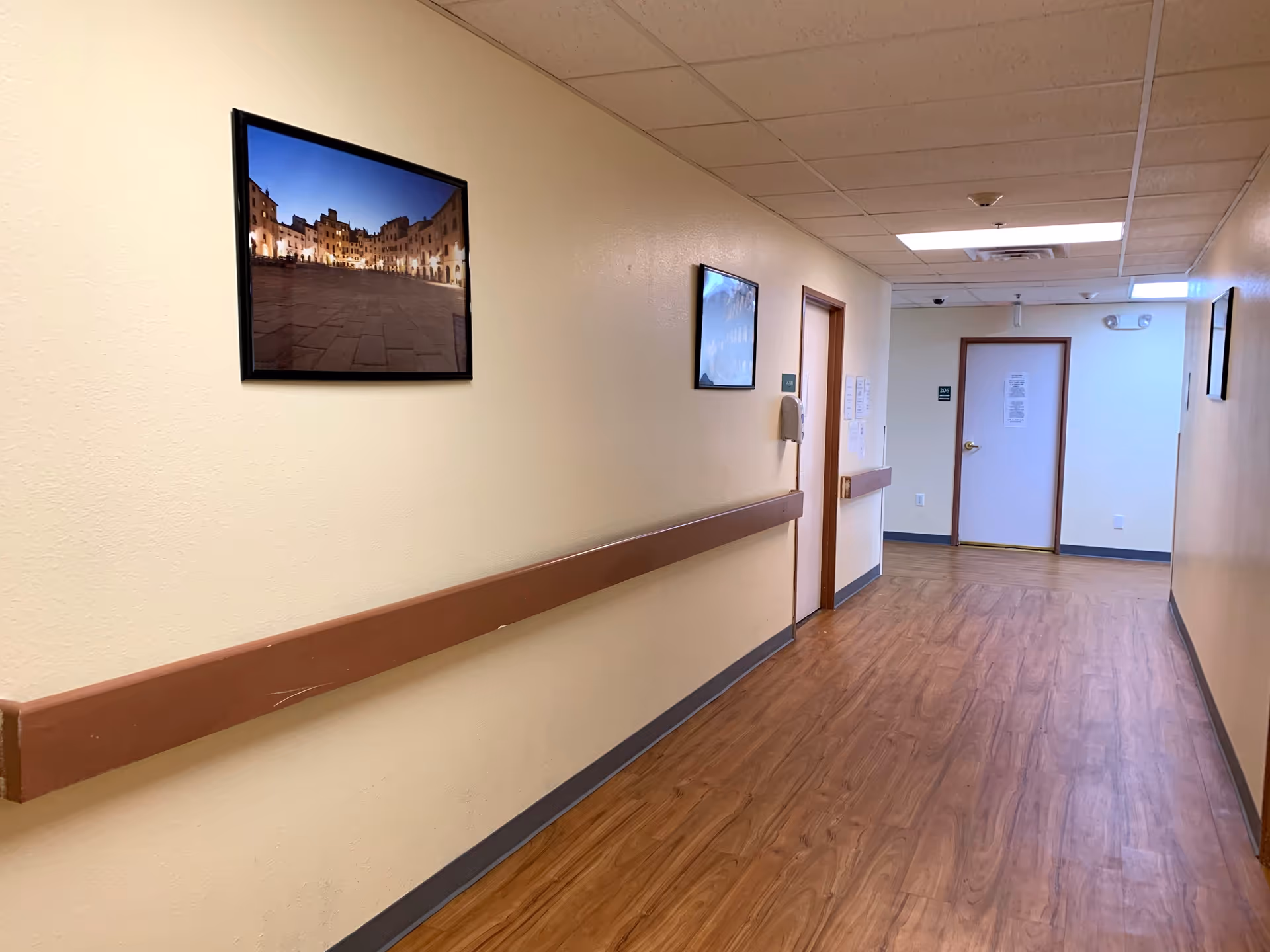 A clean, well-lit hallway in an assisted living facility with light beige walls, wood-patterned flooring, and handrails along both sides. Several framed pictures hang on the walls, and there are doors leading to rooms at the end and along the hallway.