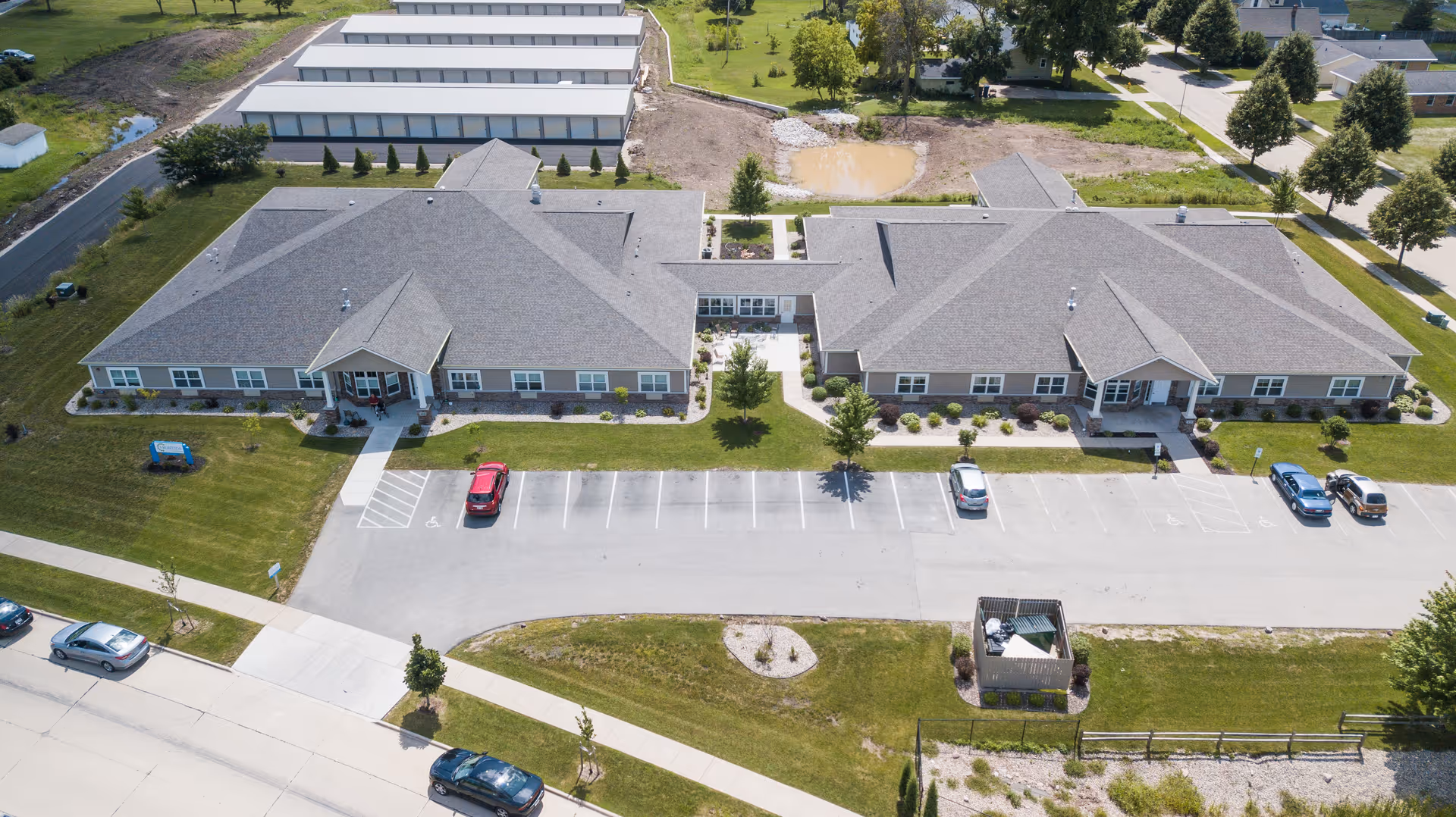 Aerial view of Frontida Assisted Living: Adelaide Place showing two large connected single-story buildings with gray roofs, surrounded by green lawns and trees. There is a parking lot in front with several cars parked, sidewalks, and a small fenced area with outdoor seating. The background includes additional buildings and open grassy areas.