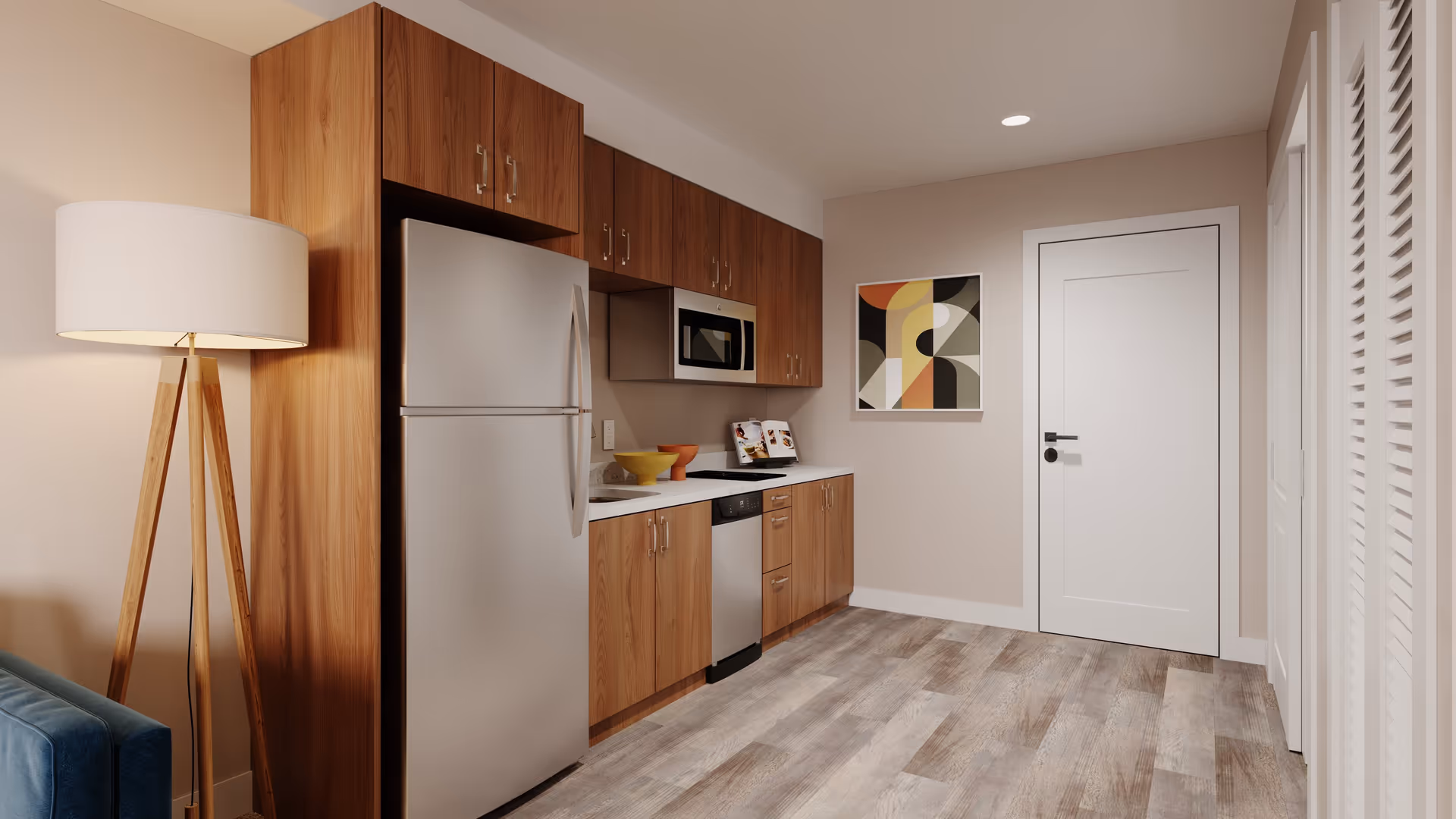 Small modern kitchenette with stainless steel refrigerator, microwave, wooden cabinets, and a floor lamp.