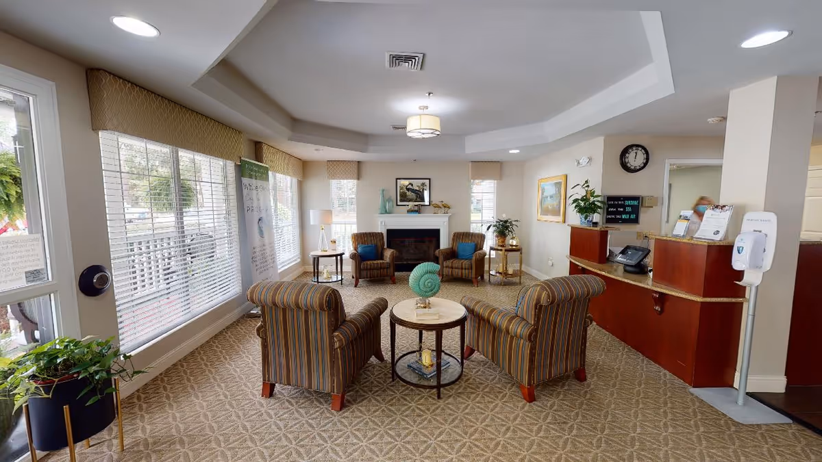 A bright and welcoming senior living facility common area with large windows covered by blinds, four striped armchairs arranged around a small round table with a decorative shell centerpiece, a fireplace with artwork above it, potted plants, a reception desk with a phone and informational materials, a wall clock, and a hand sanitizer dispenser on a stand.
