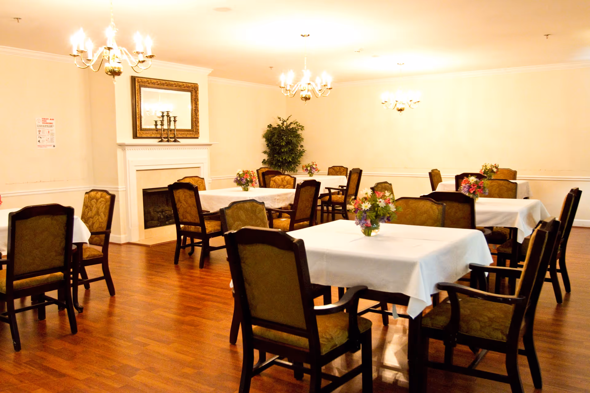 A dining room with multiple tables covered in white tablecloths, each adorned with a small vase of colorful flowers. The room has wooden floors, upholstered chairs, chandeliers hanging from the ceiling, a fireplace with a large framed mirror above it, and a potted plant in the corner.
