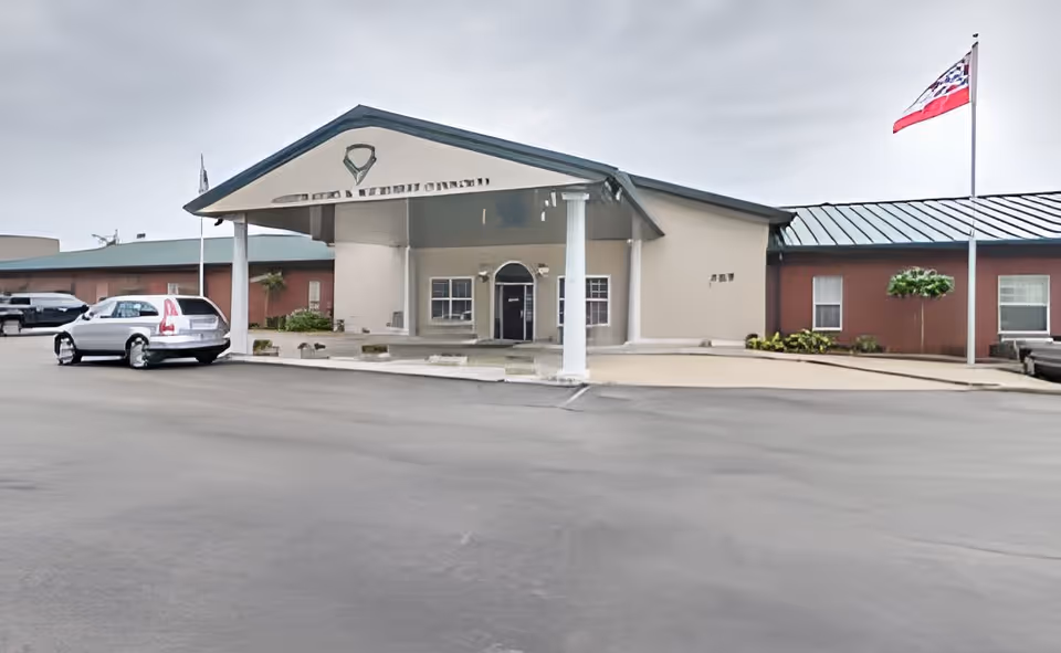 Front entrance of a single-story assisted living building with a covered porte-cochère, parked cars, and a flagpole.