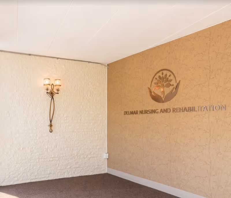 Interior corner of a room with textured beige walls, featuring a decorative wall sconce with two lights on the left wall and a metallic sign on the right wall that reads 'DELMAR NURSING AND REHABILITATION' with a logo of two hands holding a stylized tree above the text.