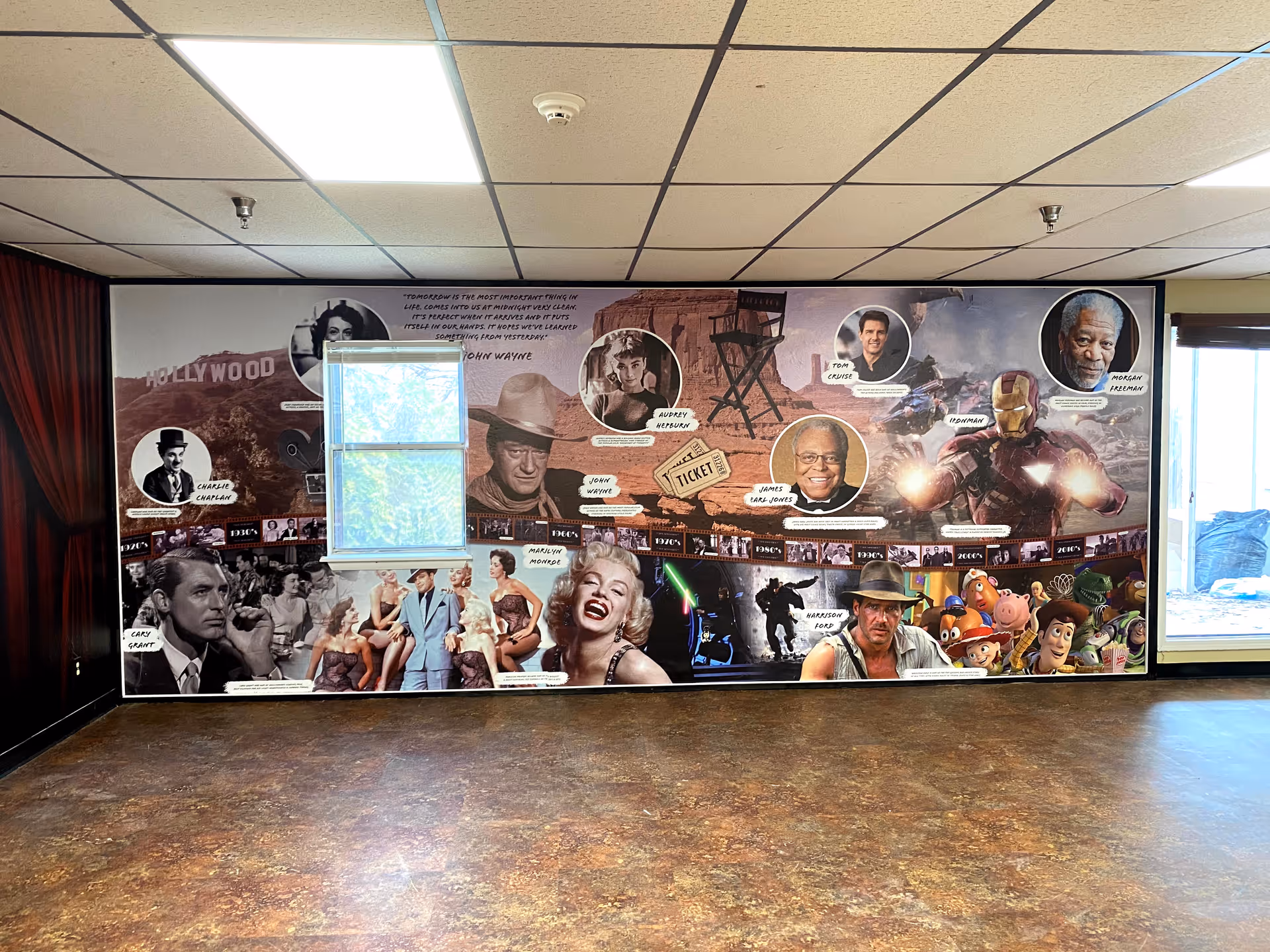 Empty interior room with a large Hollywood-themed mural covering one wall, a small window set in the mural, and a brown linoleum floor.