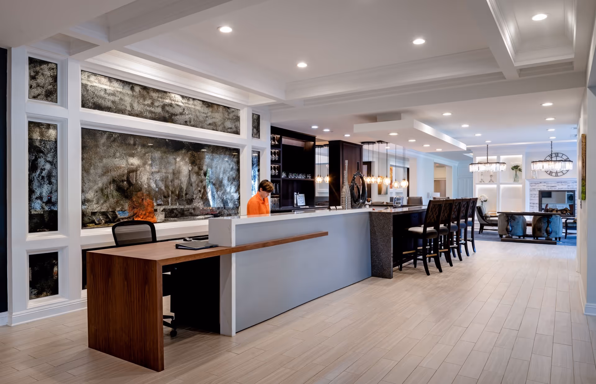 A modern, spacious interior area featuring a long reception desk with a wooden extension and a person seated behind it. The area includes a bar counter with high chairs, pendant lighting, and a lounge area with armchairs and a fireplace in the background. The space is well-lit with recessed ceiling lights and decorated with large mirrors and contemporary furnishings.