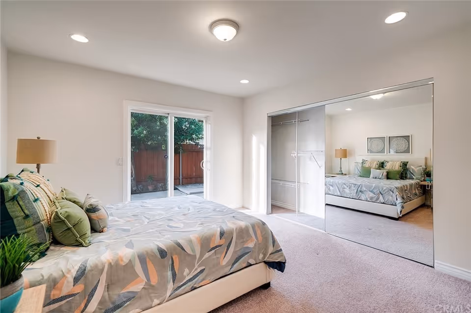 A bright bedroom with a large bed covered in a patterned comforter and multiple pillows. There is a bedside table with a lamp and a small plant. The room has a sliding glass door leading outside and a large closet with mirrored sliding doors reflecting the bed and lamp.