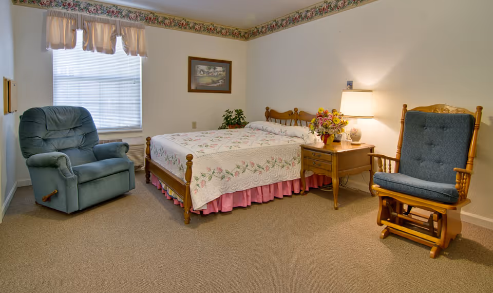 A cozy senior living bedroom with a wooden bed covered in a floral quilt, a wooden nightstand with a lamp and a flower arrangement, a blue cushioned recliner chair near a window with blinds and a valance, and a wooden rocking chair with blue cushions on a carpeted floor.