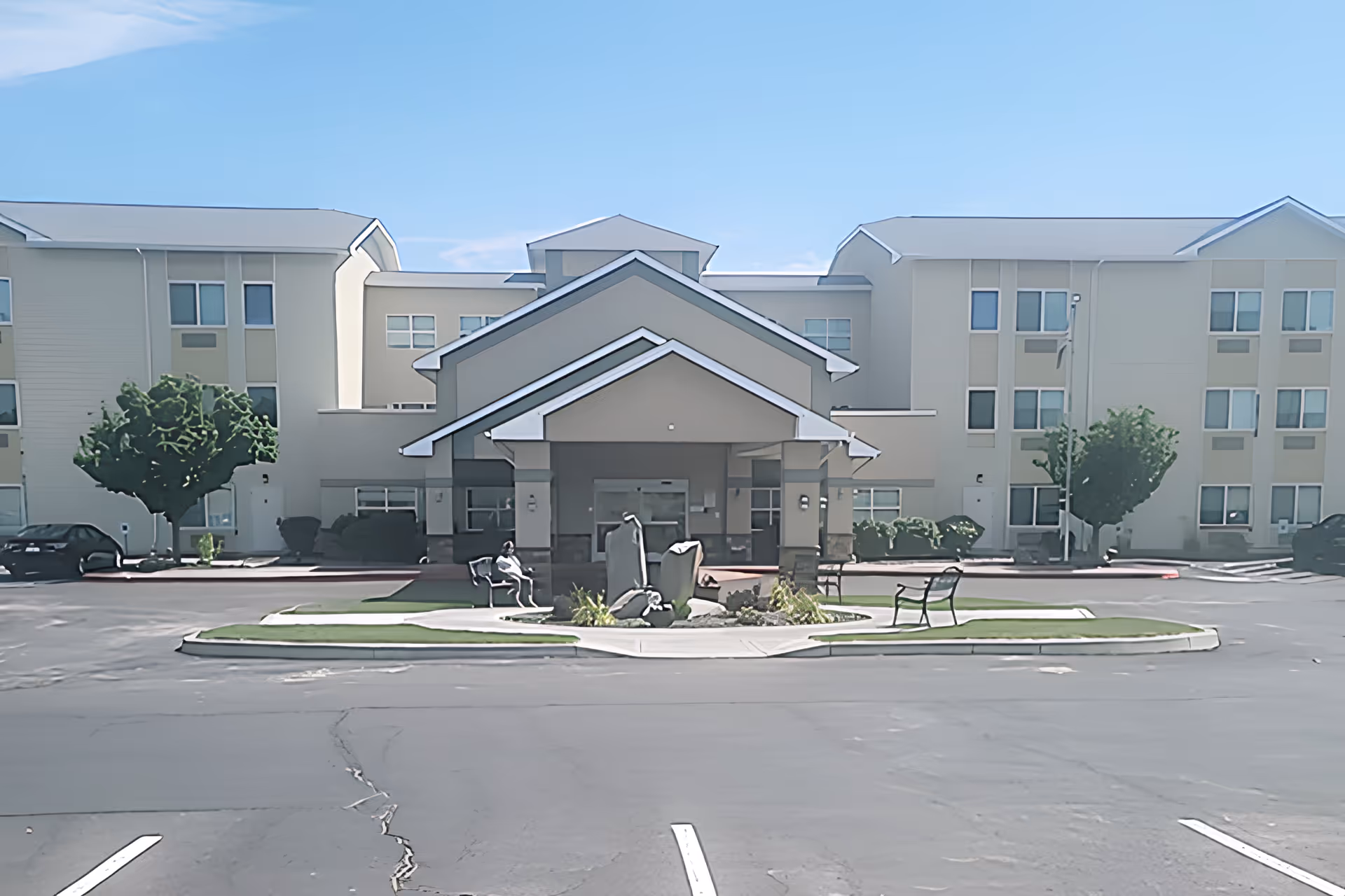 Front exterior view of a three-story senior living facility with a covered entrance, benches, and landscaped area with rocks and plants in front. The building has multiple windows and a clear blue sky above.