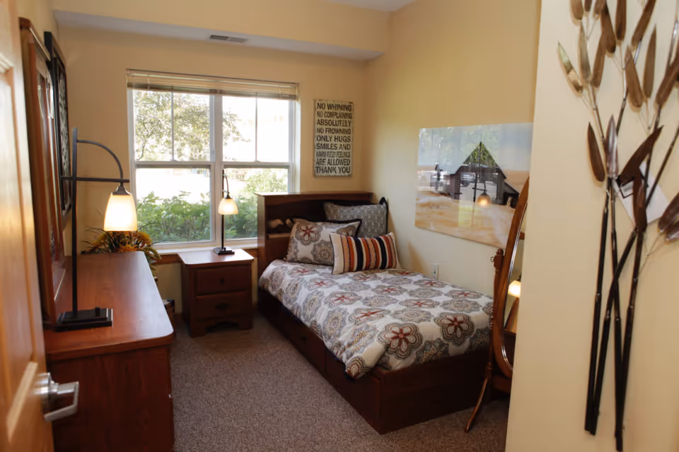 A small, cozy bedroom with a single bed featuring a floral patterned bedspread and multiple decorative pillows. There is a wooden nightstand with a lamp next to the bed, and a wooden dresser with another lamp on the opposite side. A large window lets in natural light, and the walls are painted a soft yellow. Decorative wall art and a standing mirror are also visible.