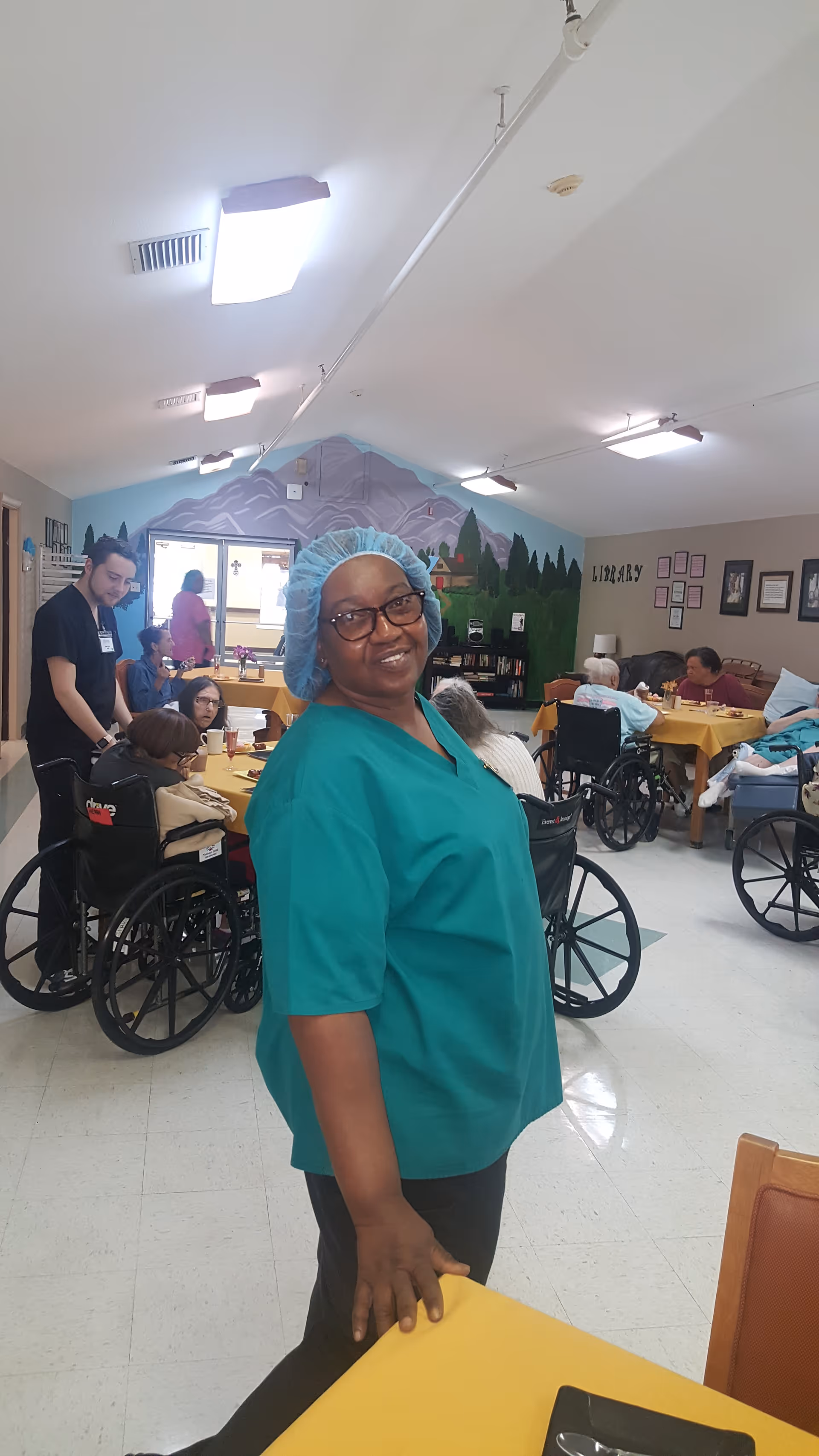 A smiling caregiver in green scrubs and a hairnet stands in a communal dining/activity room with residents in wheelchairs at tables and a mountain mural on the back wall.