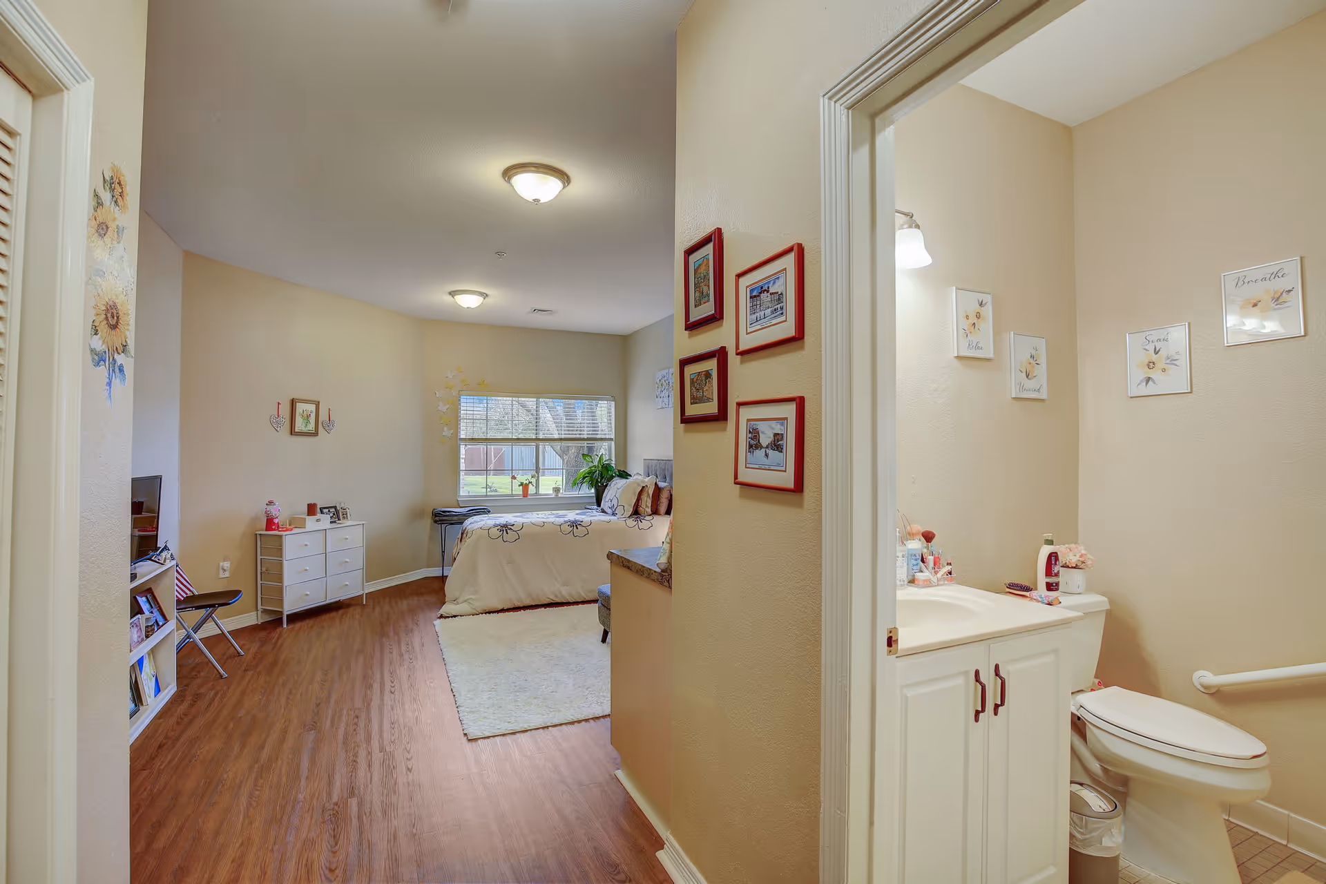 View of a senior living facility room showing a bedroom with a bed, dresser, TV, and window with a garden view. To the right, there is a bathroom with a toilet, sink, and wall decorations. The room has wooden flooring and beige walls with framed pictures and floral decorations.