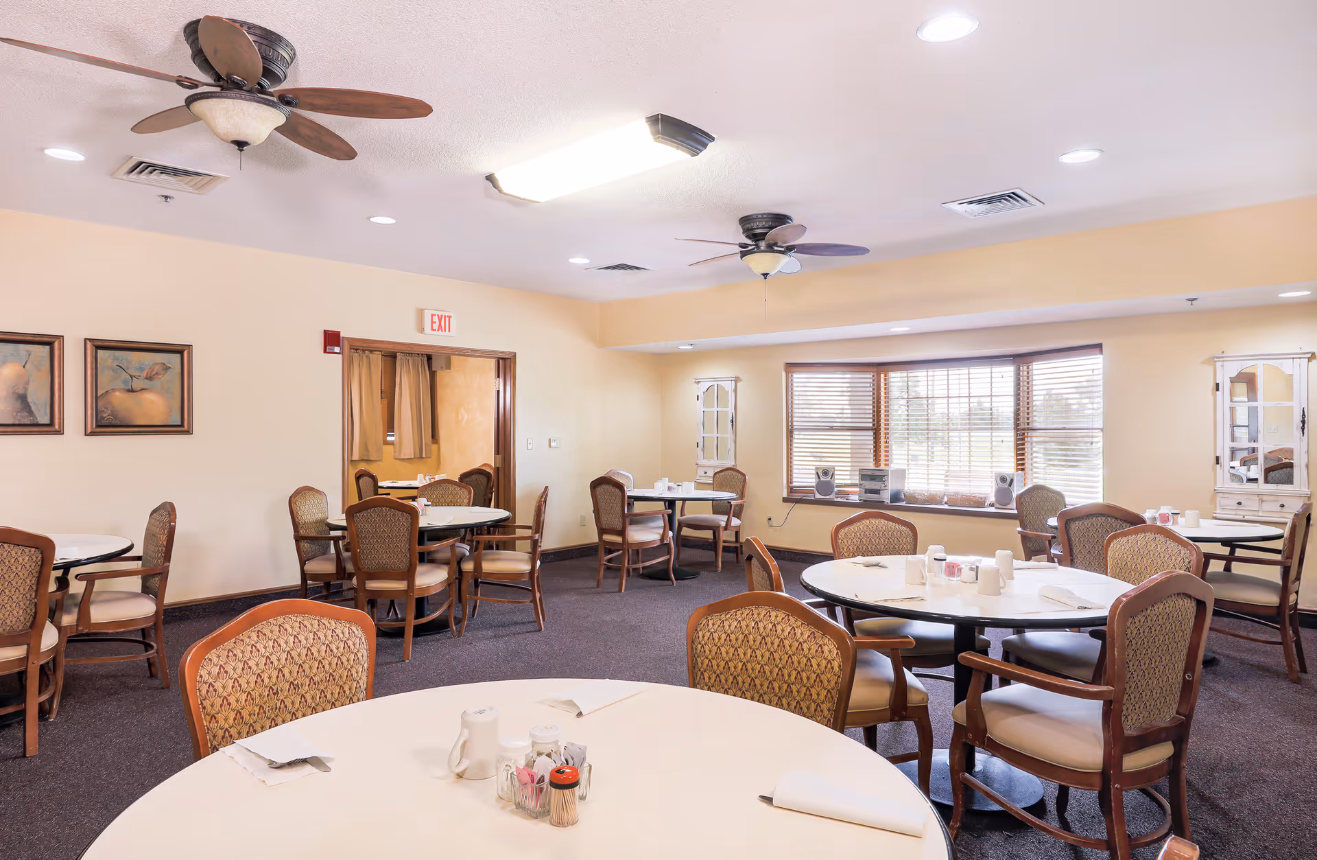 A bright dining room with several round tables and cushioned chairs arranged neatly. The tables are set with white napkins, cups, and condiment holders. The room has beige walls, carpeted floors, ceiling fans, recessed lighting, and a large window with wooden blinds letting in natural light. There are framed pictures on one wall and two white cabinets near the window.