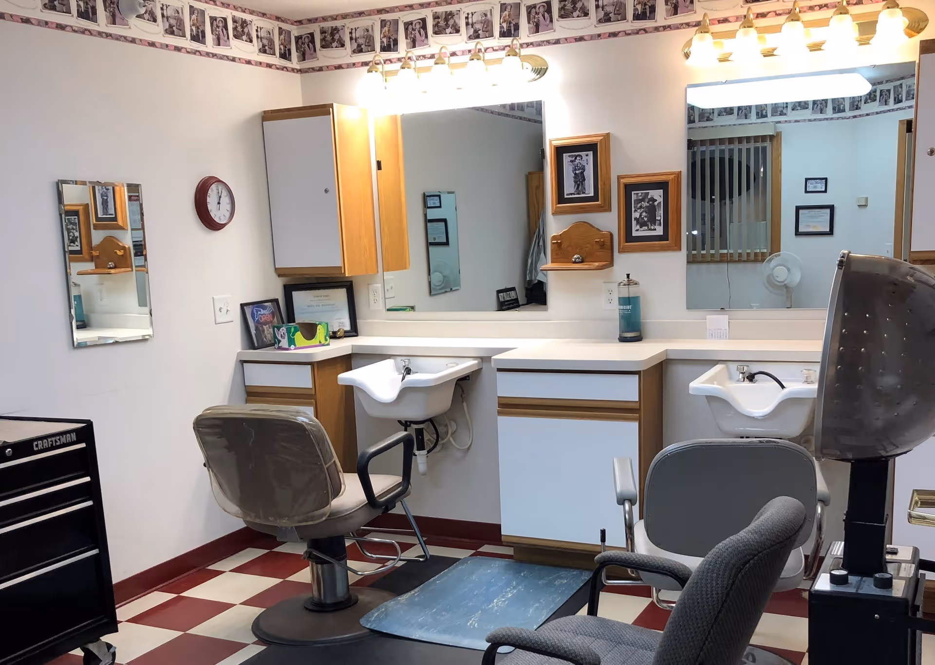 Interior of a hair salon area with two salon chairs in front of sinks and large mirrors. The room has checkered red and white flooring, wooden cabinets, framed black and white photos on the wall, and a clock. There is a hair dryer chair on the right side and a black Craftsman tool chest on the left.