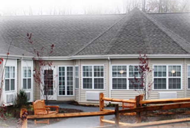 Exterior front of a single-story assisted living building with multiple large windows, a pitched shingled roof, and wooden fence and benches in the foreground.