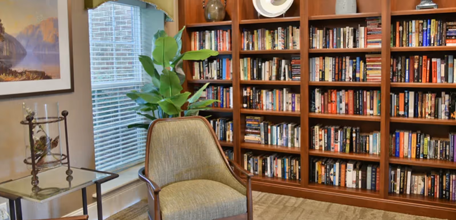 Cozy reading area with an upholstered armchair, side table, potted plant and a wall of wooden bookshelves filled with books.