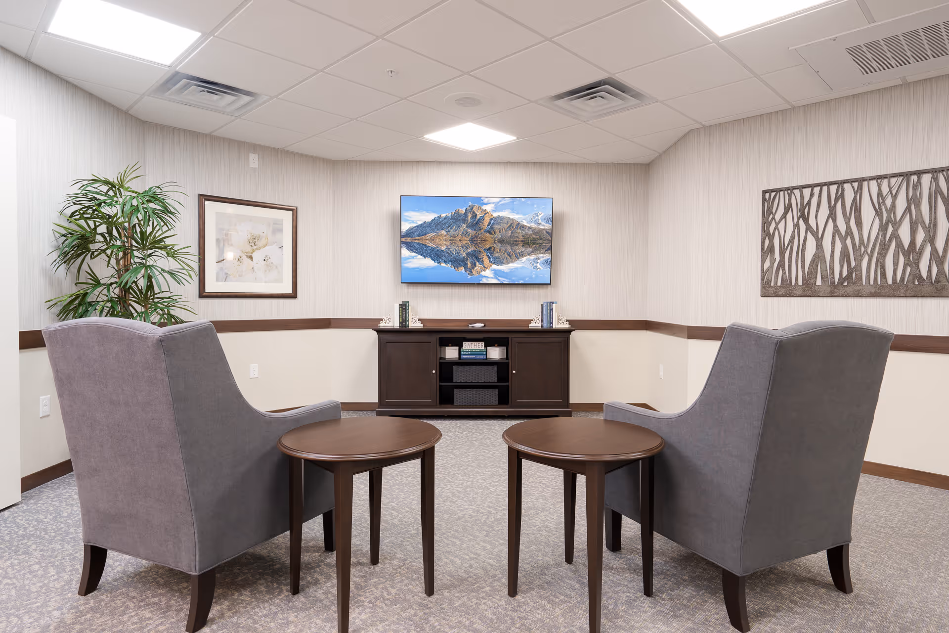 A cozy sitting area in a senior living facility with two gray armchairs facing a wall-mounted flat-screen TV displaying a mountain landscape. Between the chairs are two small round wooden tables. The room has light-colored walls with a framed picture and a decorative metal wall art. A tall green plant is placed in the corner.