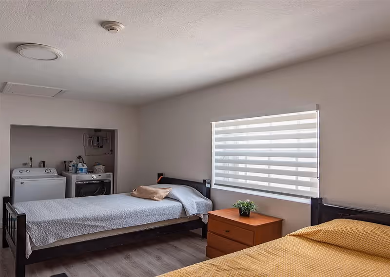 A room with two single beds, one with a gray bedspread and the other with a yellow bedspread. Between the beds is a wooden nightstand with a small green plant. There is a window with a white horizontal blind on the wall behind the nightstand. In the corner of the room, there is a laundry area with a washing machine and dryer. The floor is wooden, and the walls are light-colored.